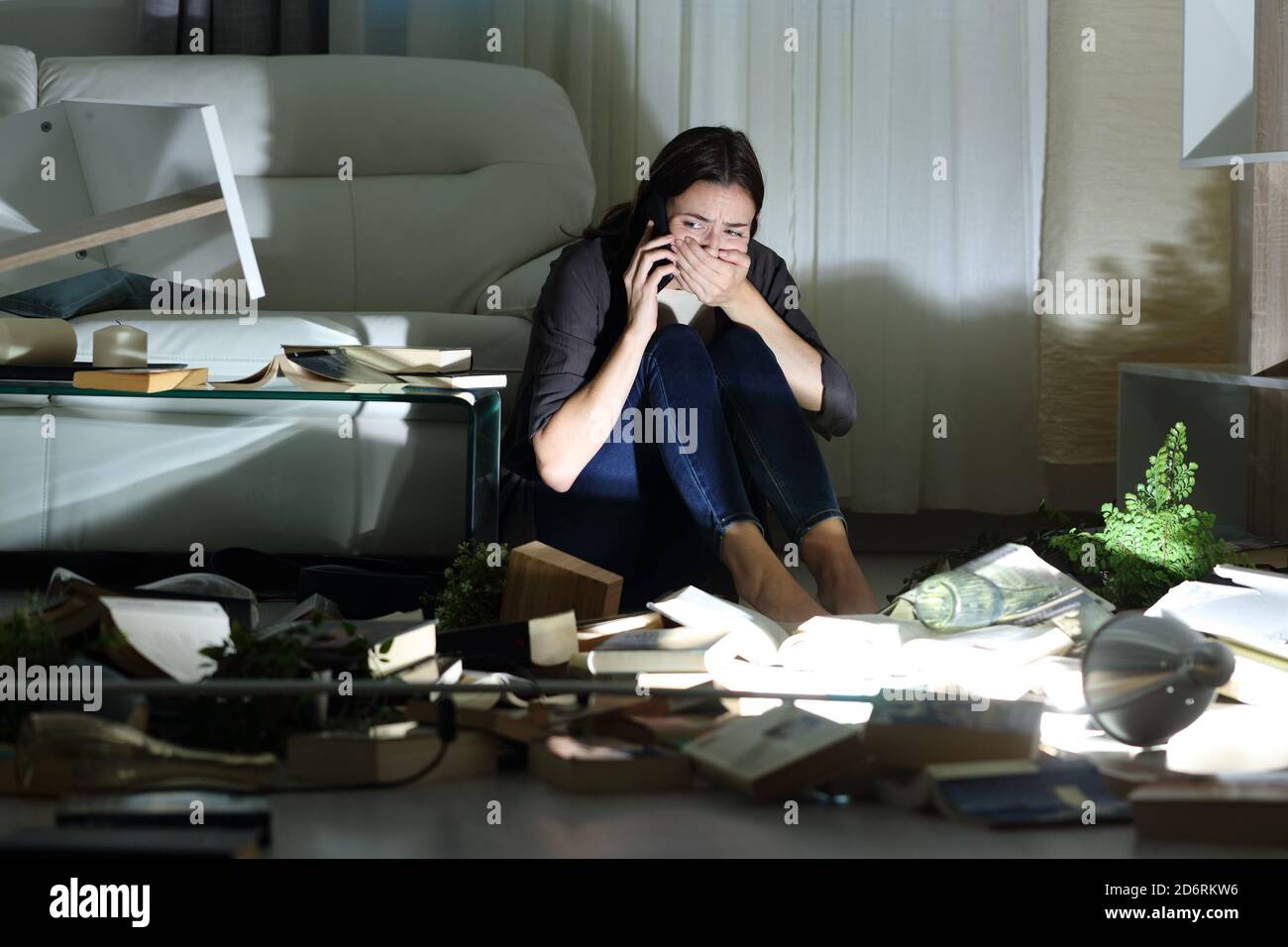 Scared home owner calling police after robbery sitting on the floor in the night with messy living room Stock Photo