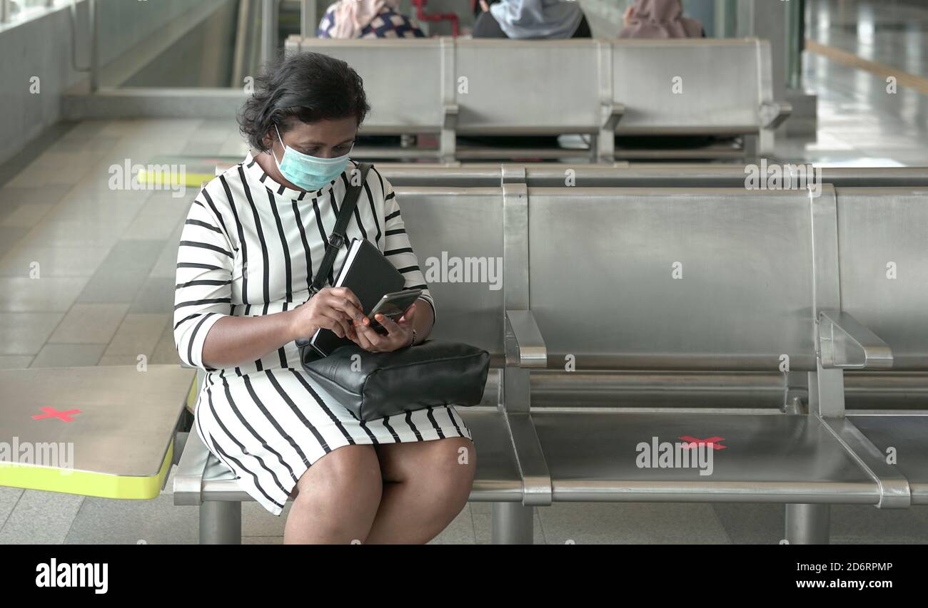 Woman wearing face mask sitting and reading her cellphone at subway station, commuters at the background. Stock Photo