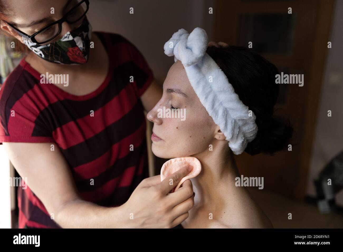 Crop anonymous makeup artist in protective mask and casual wear applying foundation with cotton pad on face of young actress with white headband Stock Photo