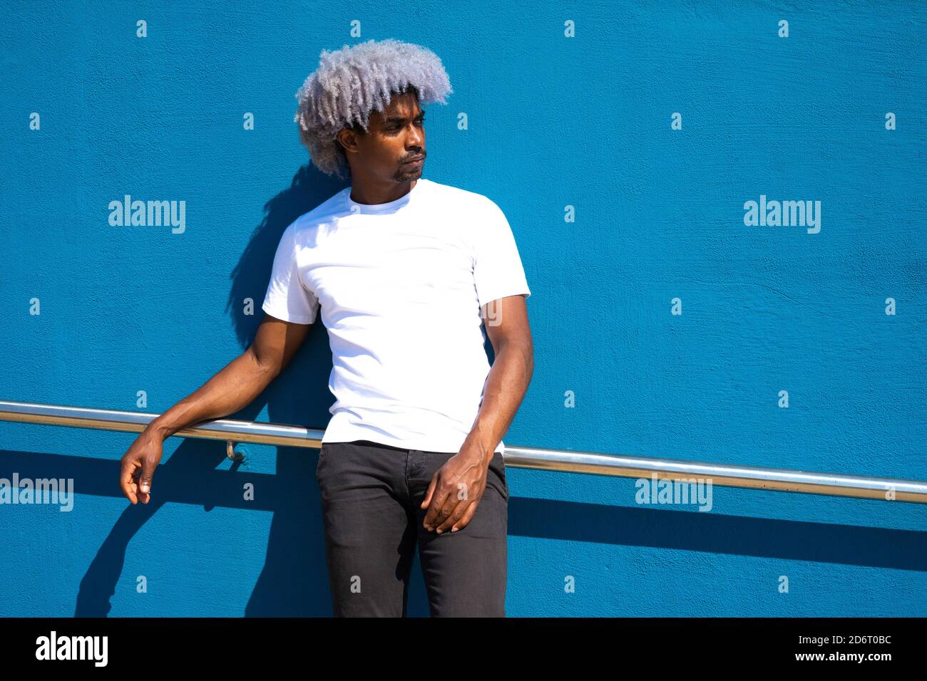 Black man with afro hair dressed in white leaning against a blue wall. Afro hair concept. Black man in white t-shirt. Stock Photo