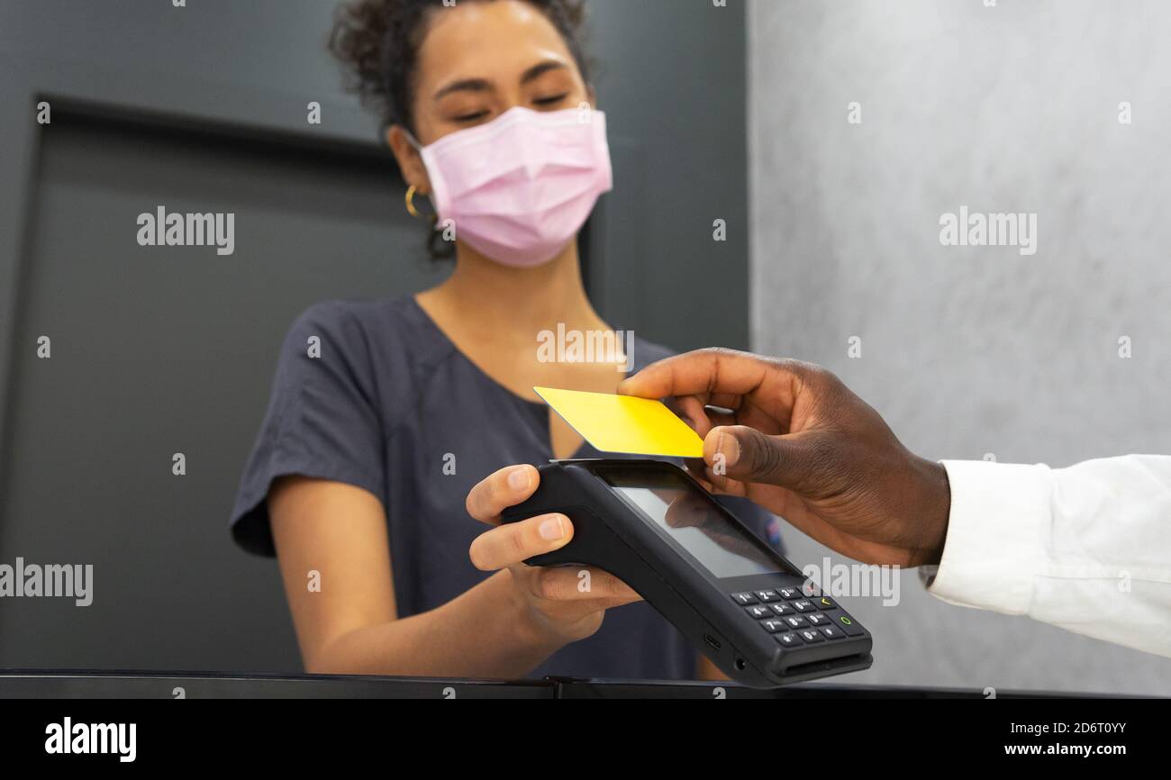 Low angle of African American female manager in medical mask receiving payment via POS terminal and plastic card from crop male patient in modern clin Stock Photo