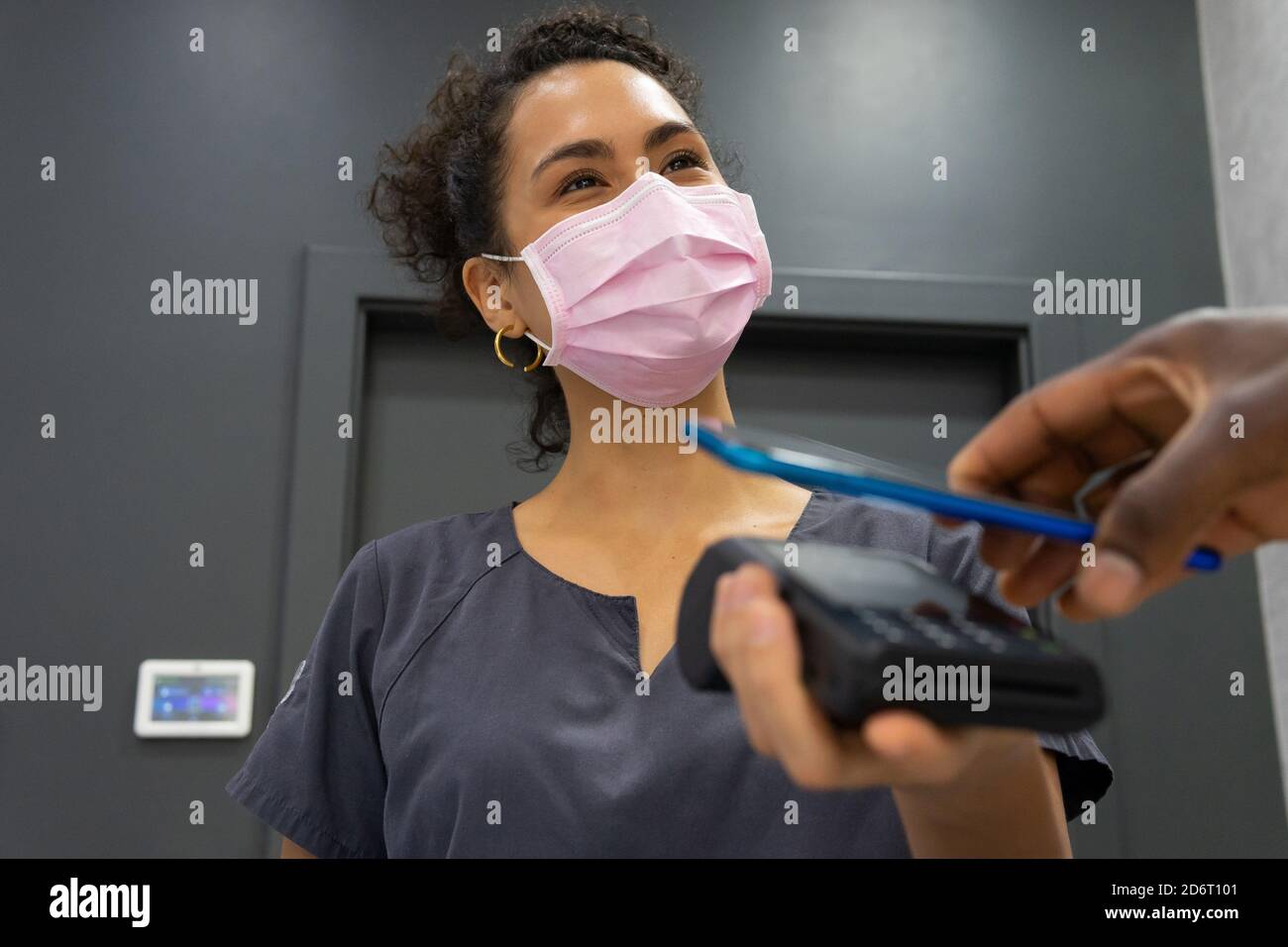 Low angle of African American female manager in medical mask receiving payment via POS terminal and mobile phone contactless payment from crop male pa Stock Photo