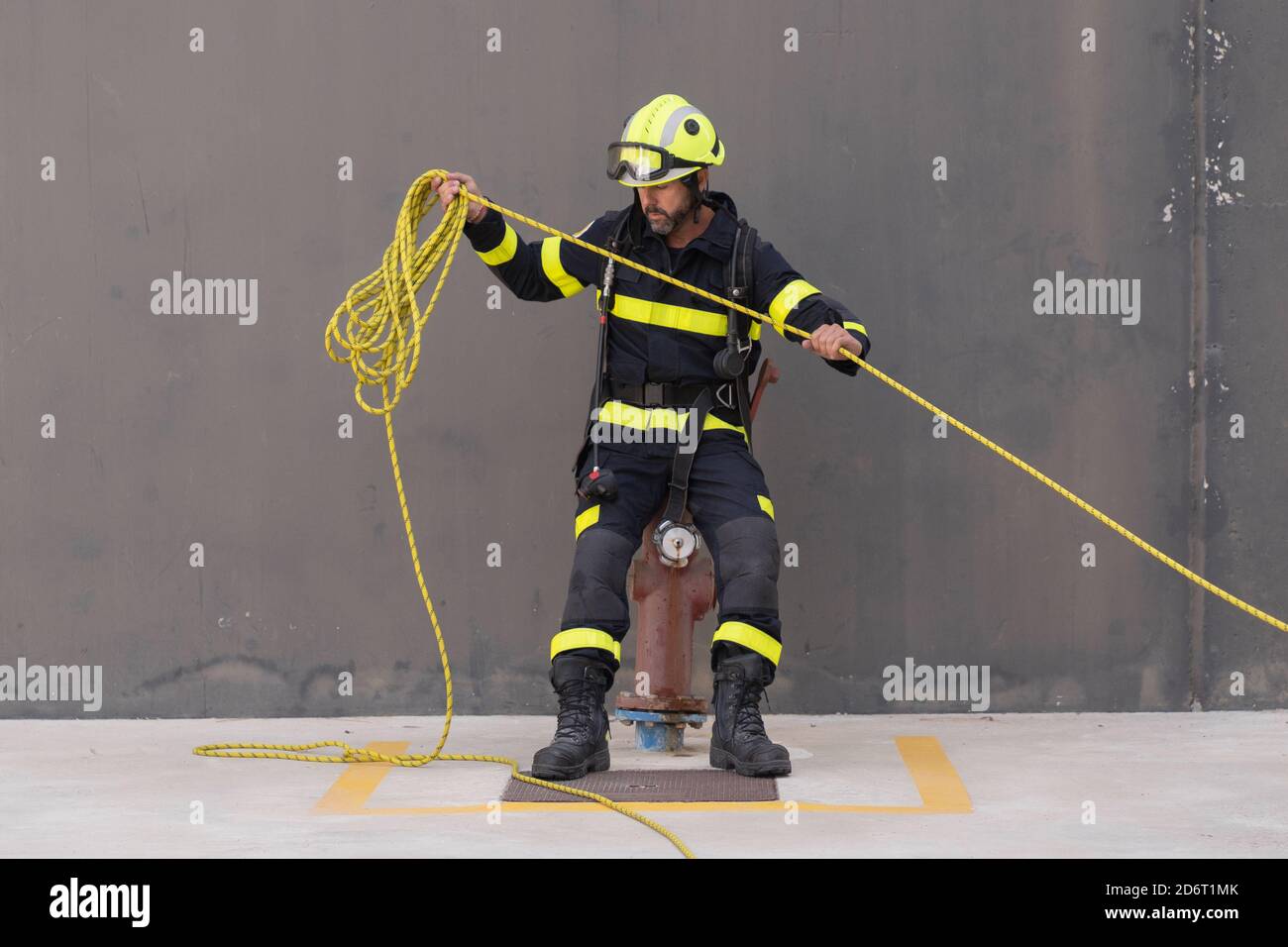 Bearded firefighter in protective hardhat and bright uniform standing on cement floor with rope during routine practices at work Stock Photo
