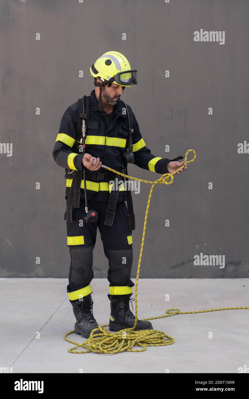 Bearded firefighter in protective hardhat and bright uniform standing on cement floor with rope during routine practices at work Stock Photo