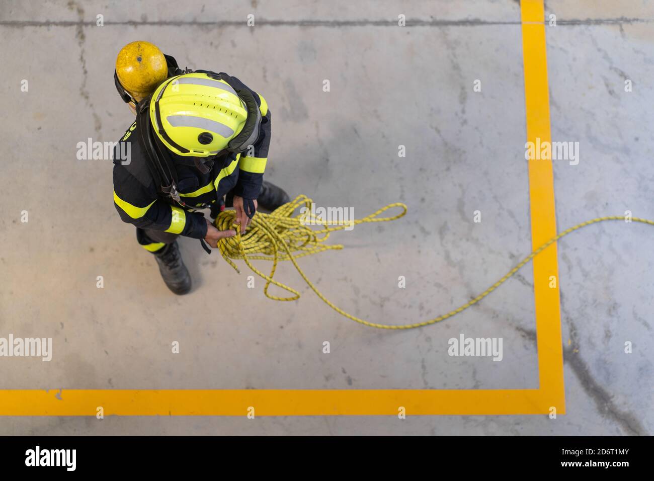 Top view of unrecognizable firefighter in protective hardhat and bright uniform standing on cement floor with rope during routine practices at work Stock Photo