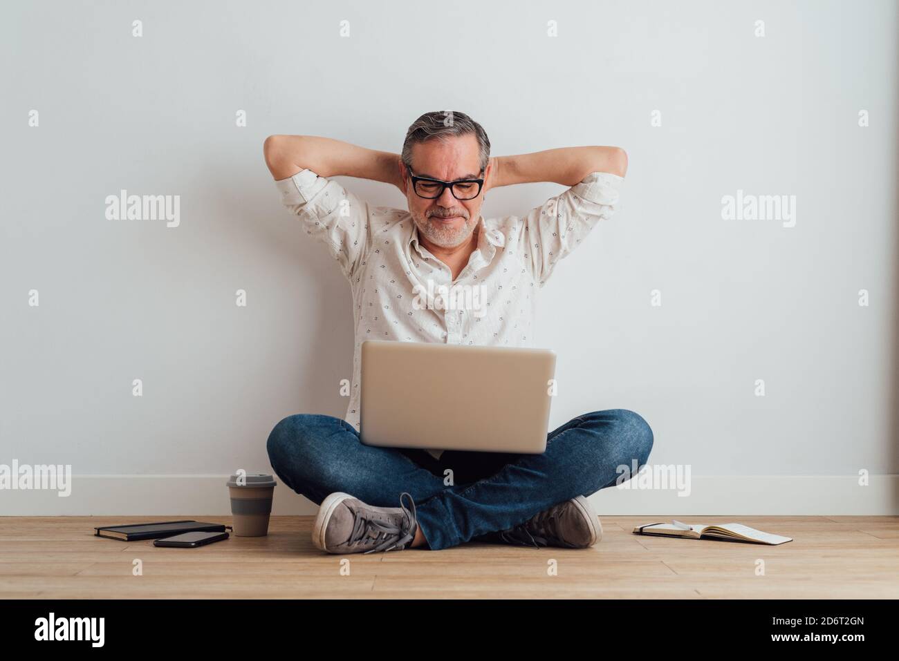 Full body delighted pleased mature male wearing casual clothes sitting on floor with contemporary laptop on laps Stock Photo