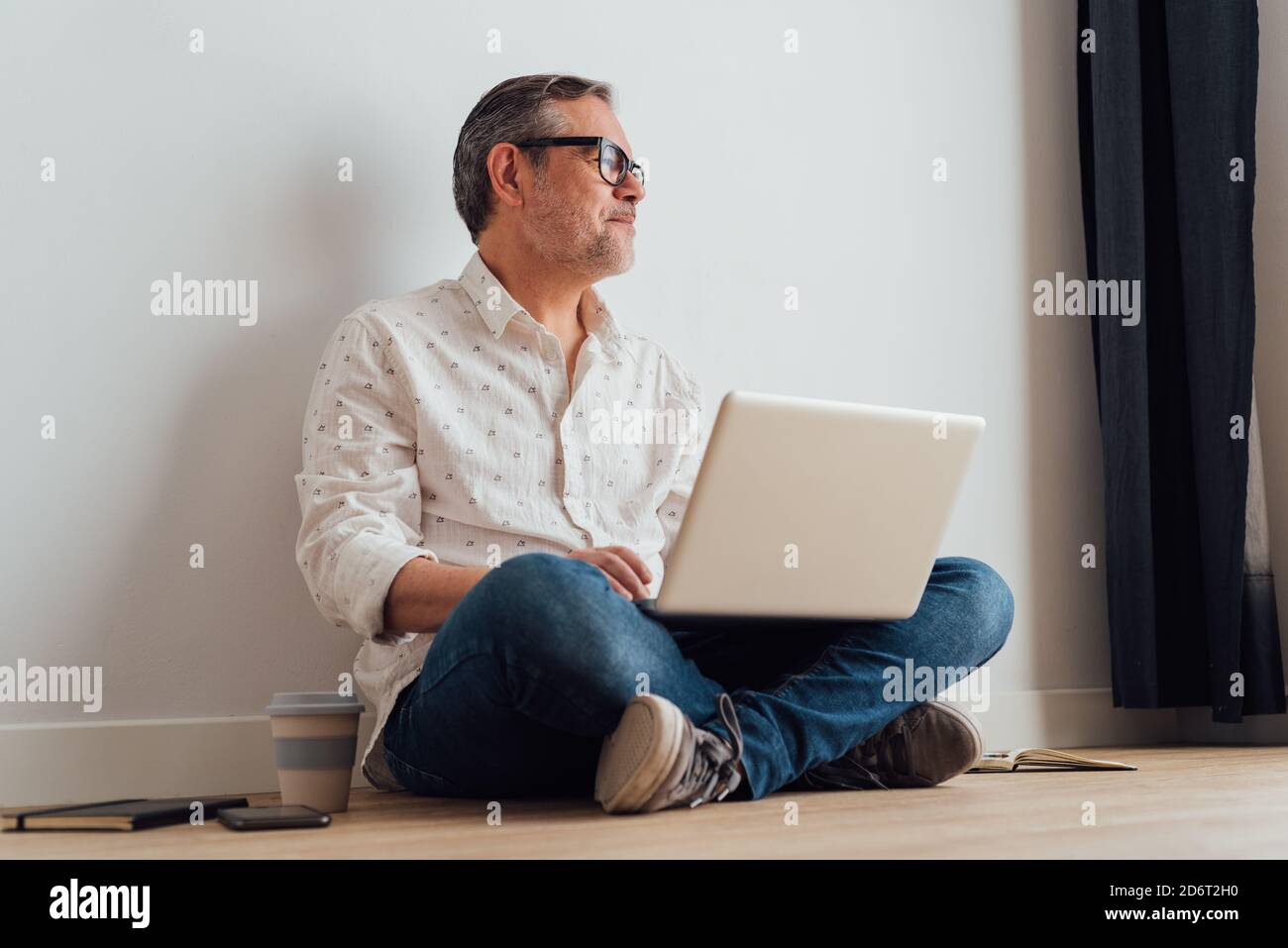 Contemplative senior male sitting with legs crossed on parquet with netbook on laps and cup of takeaway coffee in hand while looking away in thoughts Stock Photo
