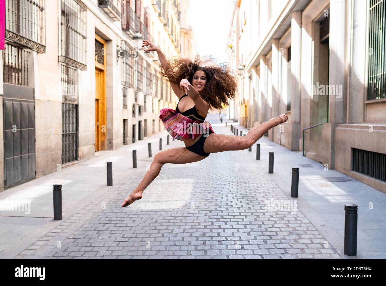 Full body of young fit ethnic female dancer with long curly hair doing grand jete position and smiling on paved street looking at camera Stock Photo