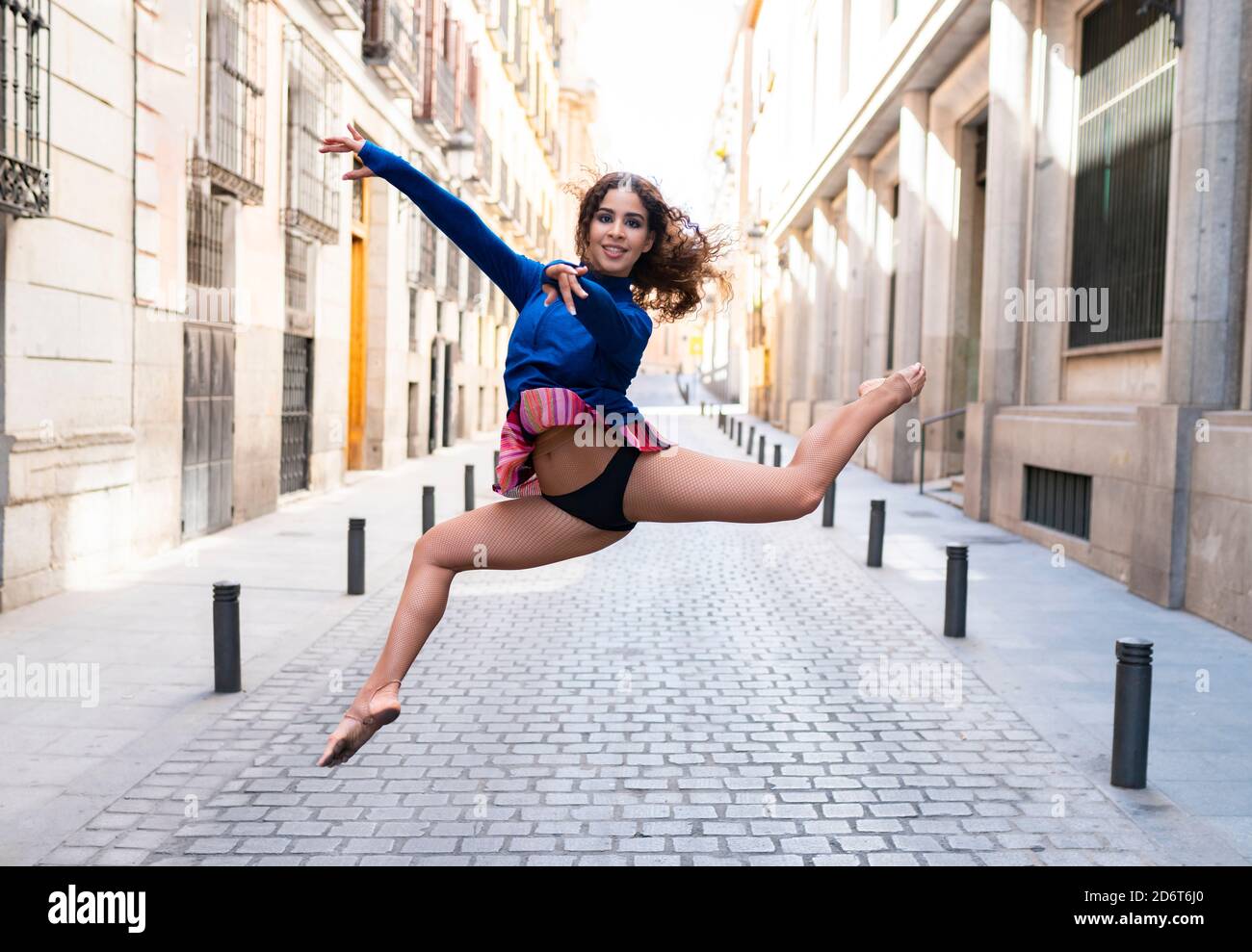 Full body of young fit ethnic female dancer with long curly hair doing grand jete position and smiling on paved street looking at camera Stock Photo