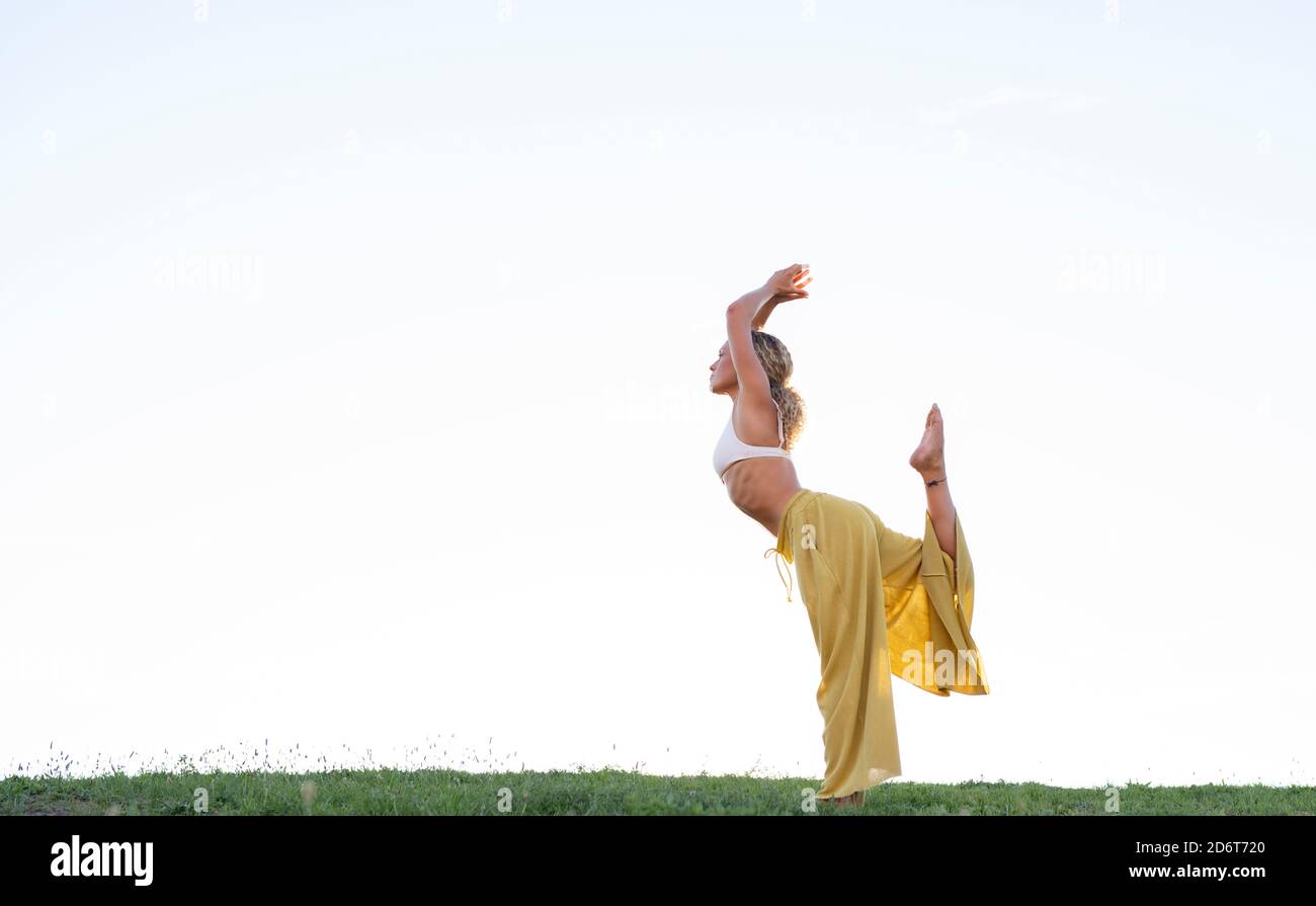 Side view full body barefoot female in bra and loose trousers performing Dancers Pose and outstretching hand with cup of coffee while practicing yoga Stock Photo