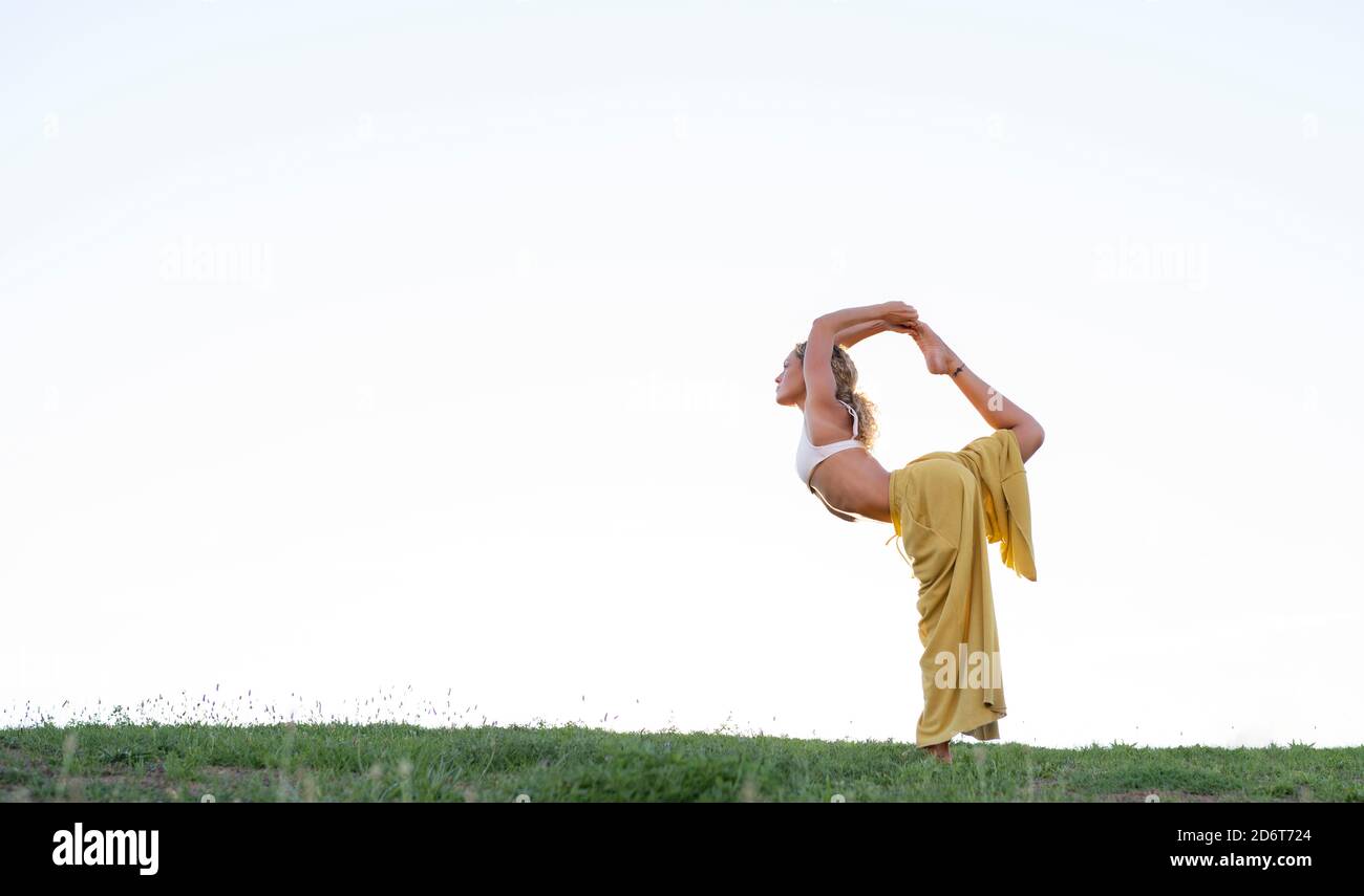 Side view full body barefoot female in bra and loose trousers performing Dancers Pose and outstretching hand with cup of coffee while practicing yoga Stock Photo