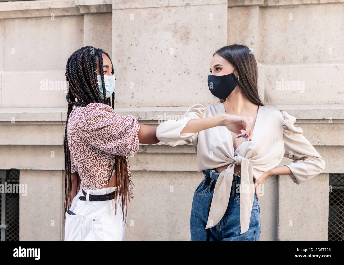 Side view of positive young multiracial female friends in trendy clothes and protective masks greeting each other with elbow bump on city street durin Stock Photo
