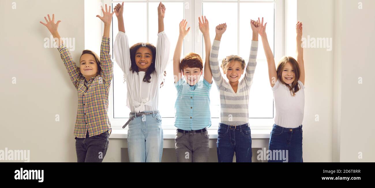 Group of happy diverse kids standing by the window with arms raised, smiling and looking at camera Stock Photo