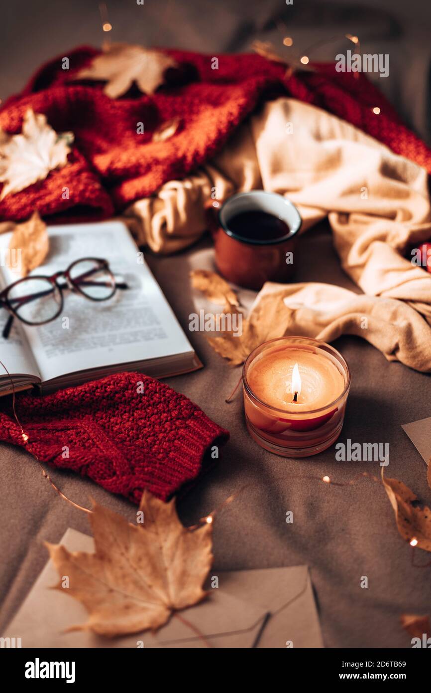 Burning candles, cup of hot tea, open book and glasses on plaid in bed. Autumn leaves and light garland decor on sofa. Autumn style. Hygge concept, vertical photo orientation Stock Photo