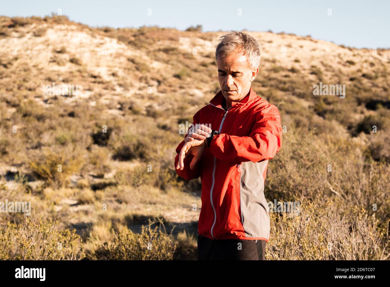 Concentrated middle aged male athlete in activewear checking time on smartwatch while standing in hilly terrain after workout Stock Photo