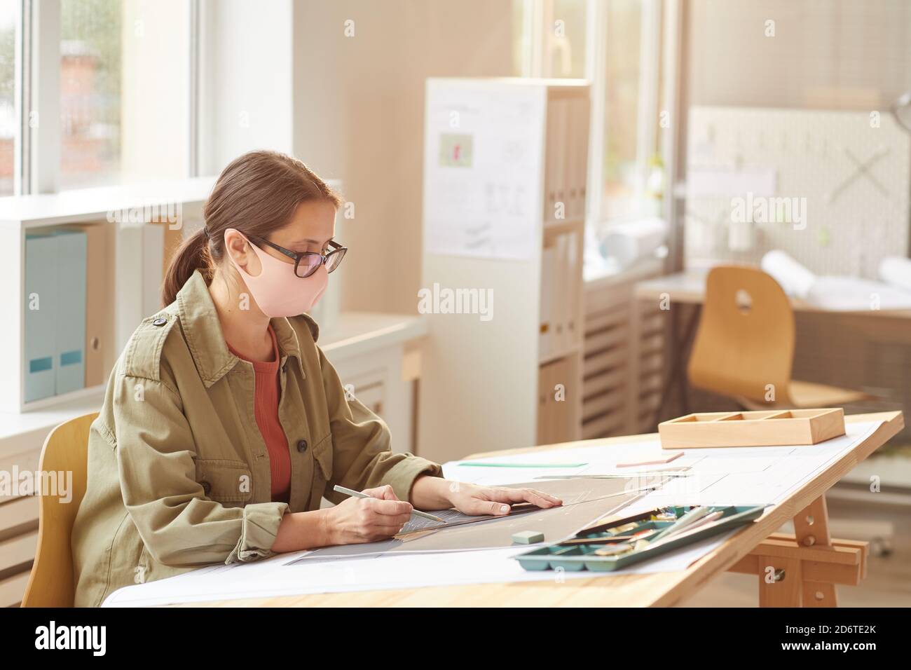 Side view portrait of female architect wearing mask while sitting at drawing desk in sunlight, copy space Stock Photo