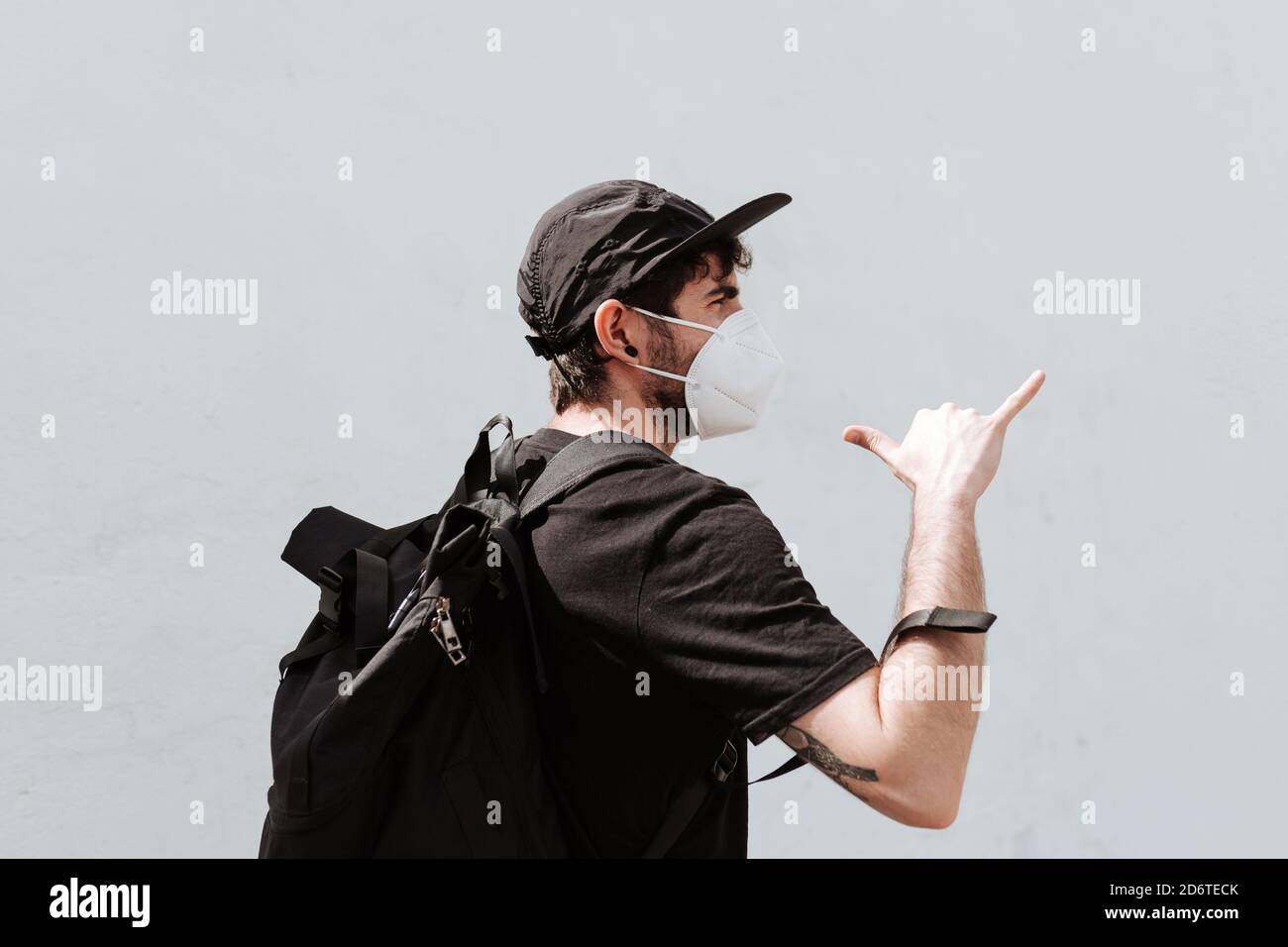 Back view of unrecognizable trendy man with backpack in respiratory mask demonstrating shaka gesture near wall while looking away during coronavirus p Stock Photo