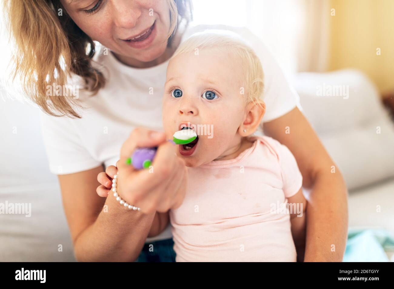 Crop smiling Woman holding cute blond haired toddler and feeding with spoon in bright room on blurred background Stock Photo