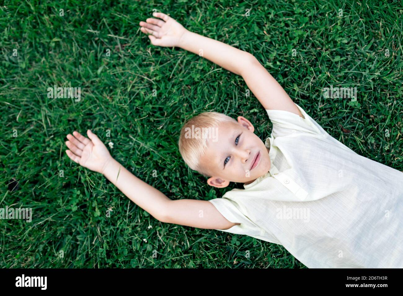 Overhead cheerful blond haired boy in white shirt looking at camera with smile while chilling on verdant grass with arms behind head Stock Photo