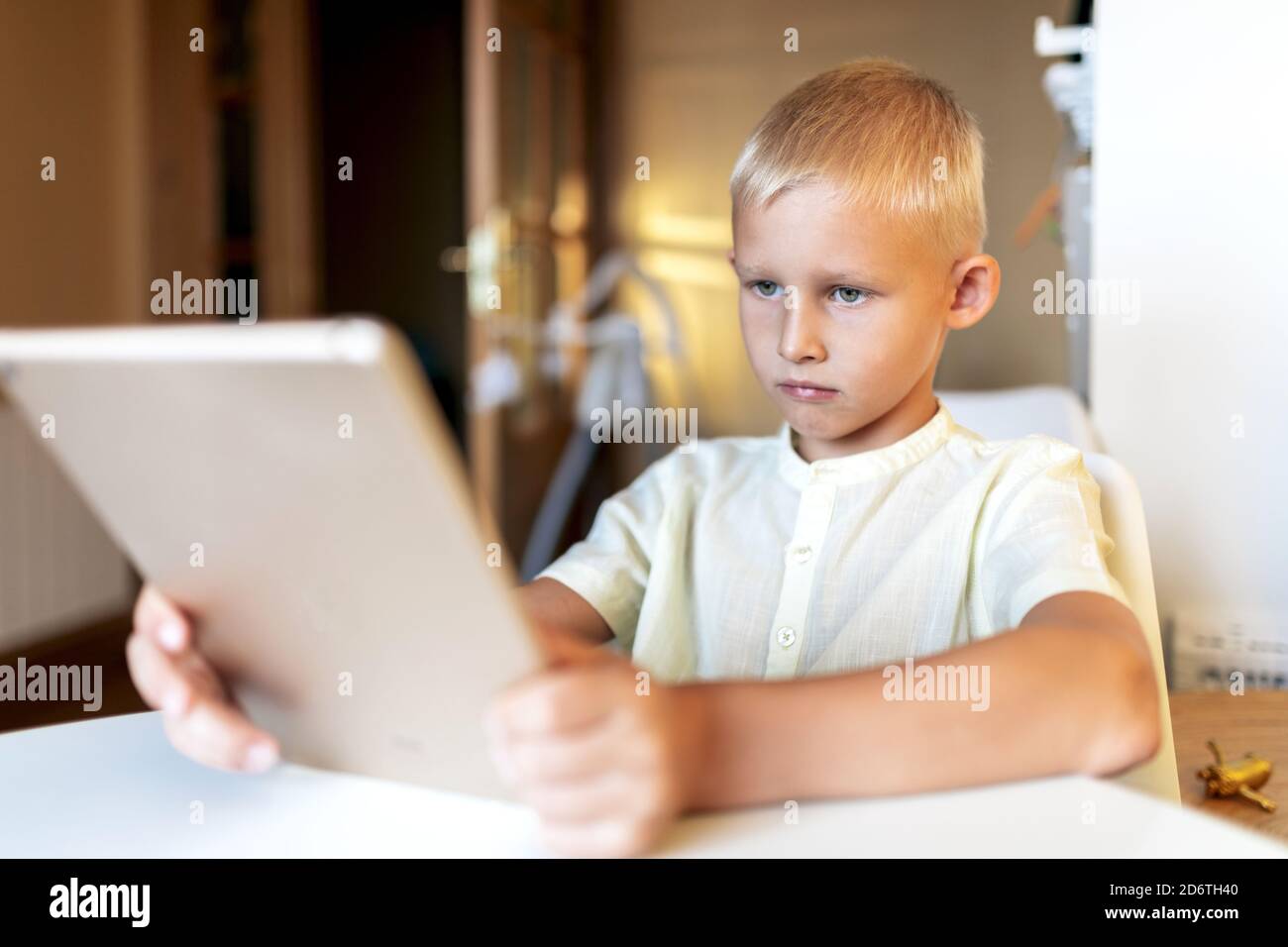 Side view of concentrated blond haired little boy in white shirt browsing internet on tablet in bright room in sunlight Stock Photo