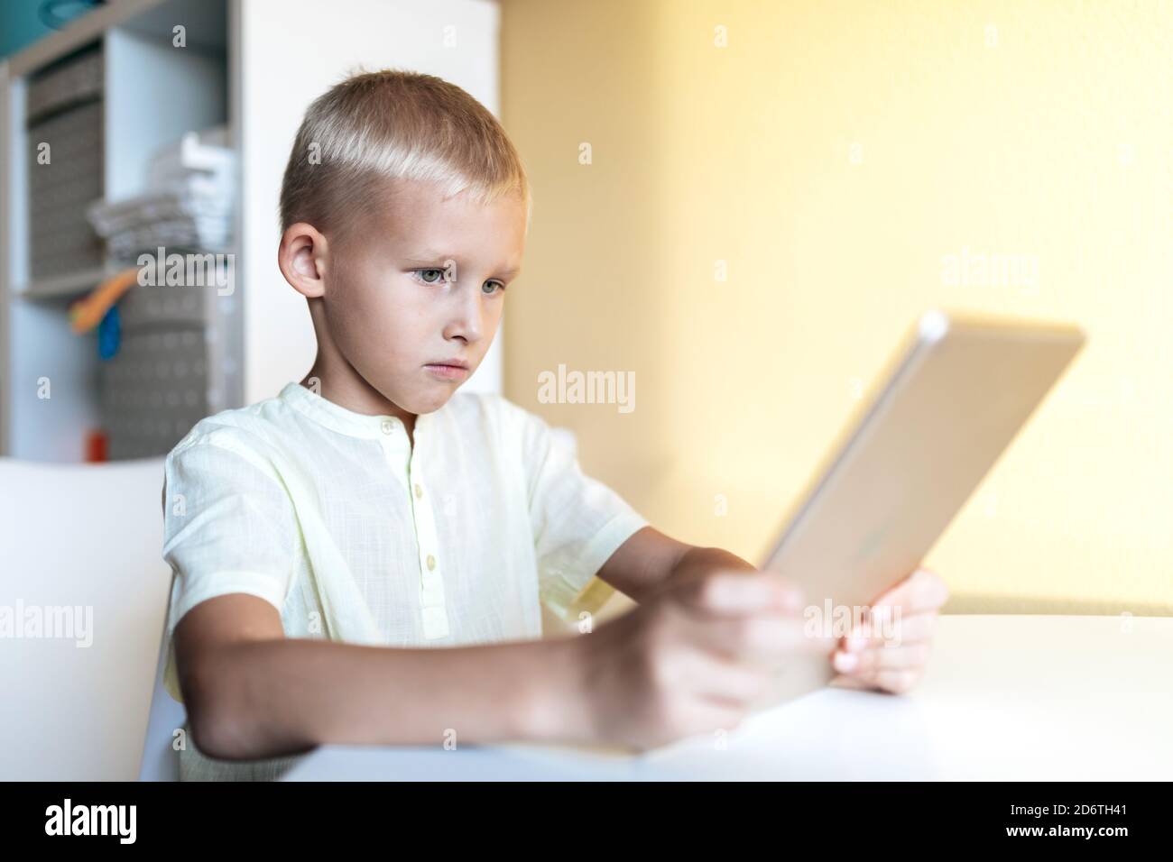 Side view of concentrated blond haired little boy in white shirt browsing internet on tablet in bright room in sunlight Stock Photo