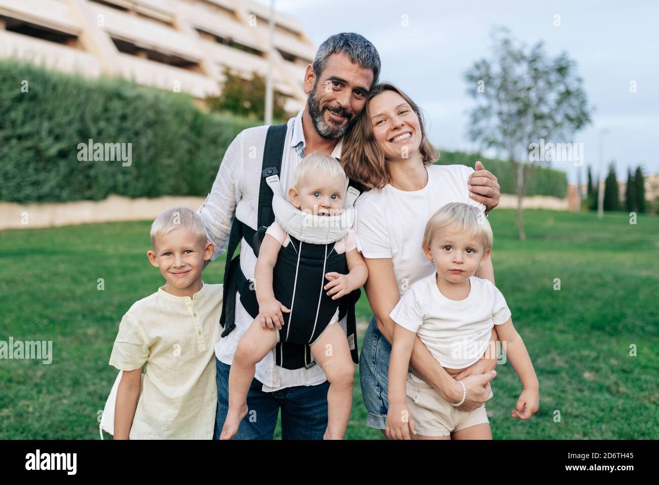 Cheerful father and mother looking at camera with smile while hugging little blond haired sons in cozy park next to building immersed in greenery Stock Photo
