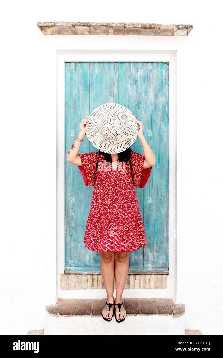 Anonymous female traveler in trendy apparel covering face with straw hat near aged building in Greece Stock Photo