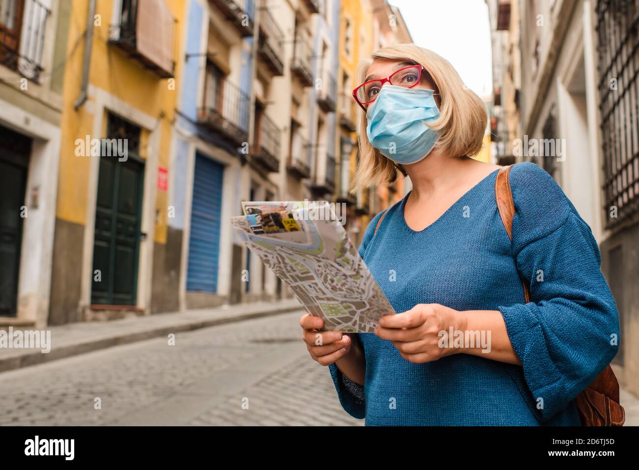Low angle of female traveler in medical mask for coronavirus prevention holding touristic map and checking route while standing on narrow paved street Stock Photo