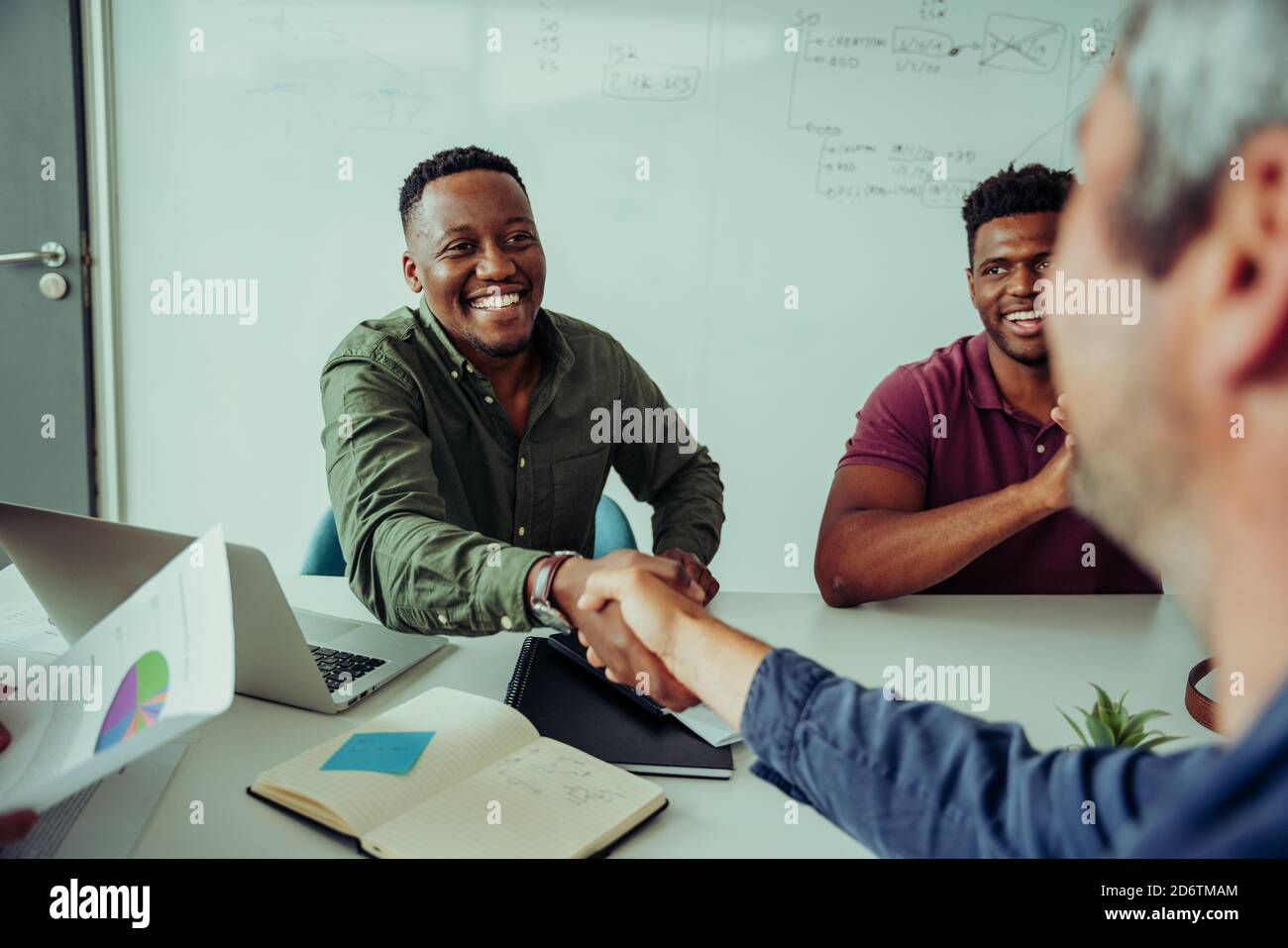 Smiling mixed race business man shaking hands with manager accepting project deal sitting in boardroom during team meeting  Stock Photo