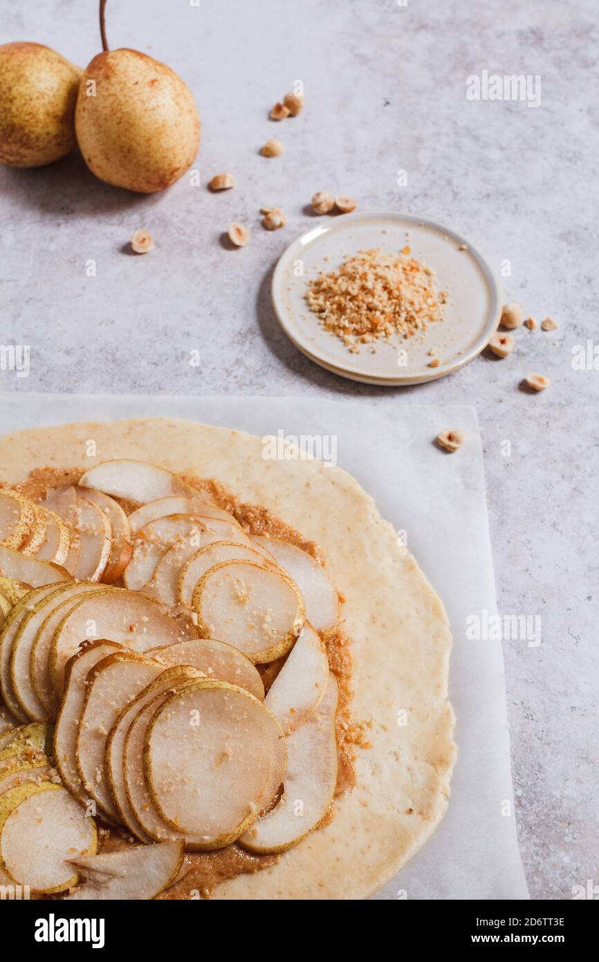 Preparation of a pear and hazelnut galette. The rolled raw dough is spread with hazelnut praline and layered with pear slices. Stock Photo