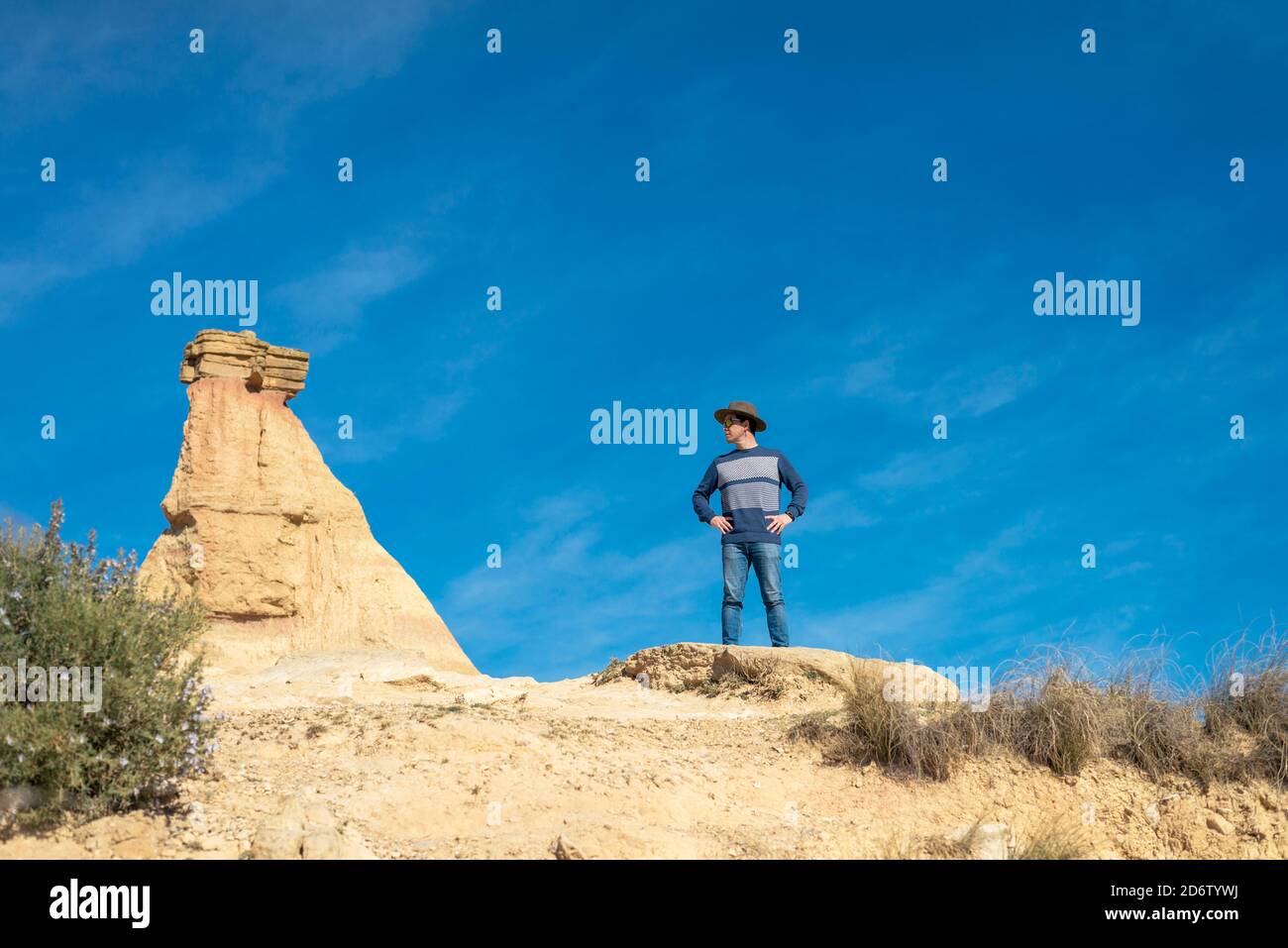 Front view of a man with hat and sunglasses standing on desert dunes against a rock peak Stock Photo