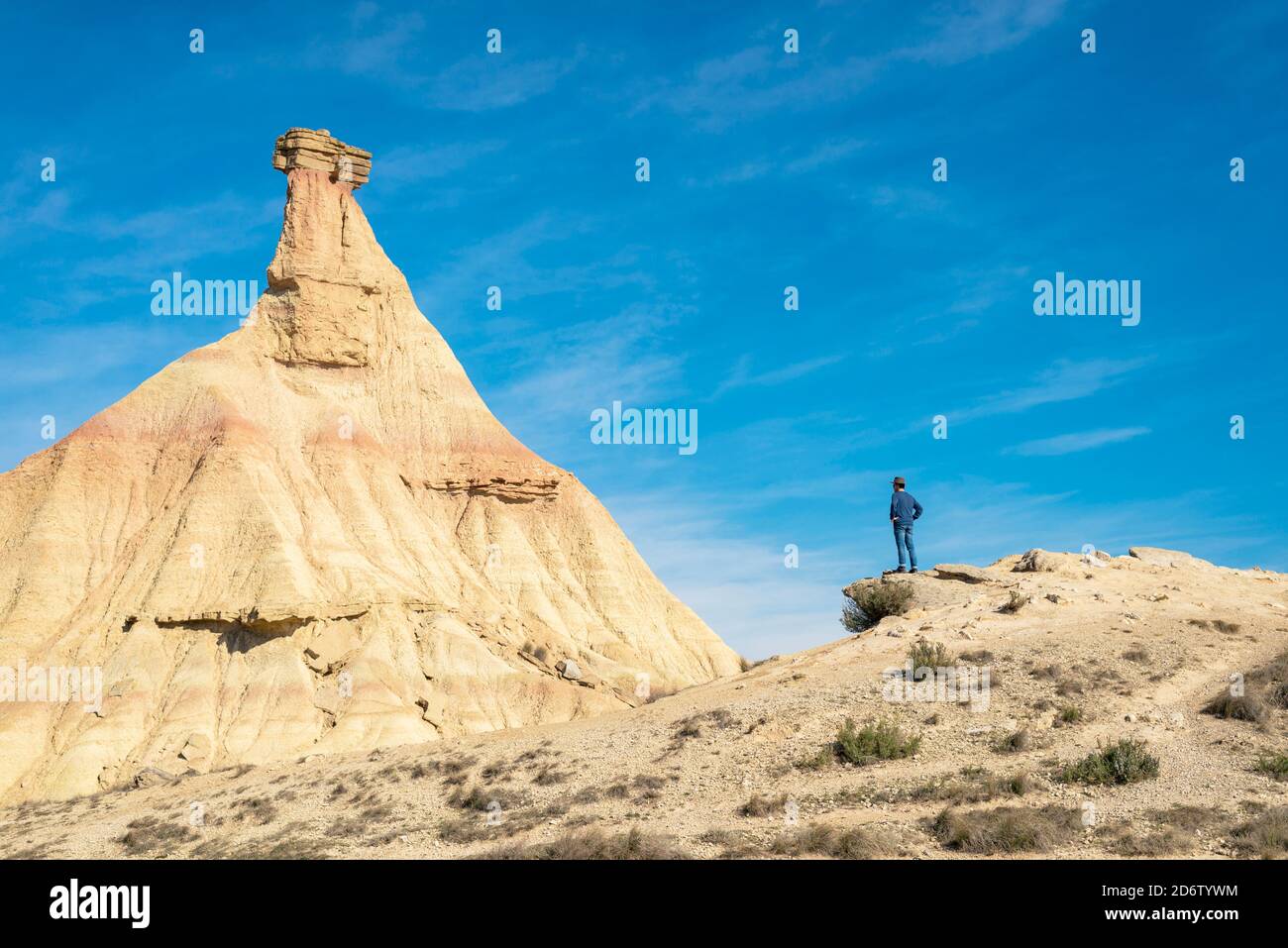 Rear view of a man with hat and sunglasses standing on desert dunes against a rock peak Stock Photo