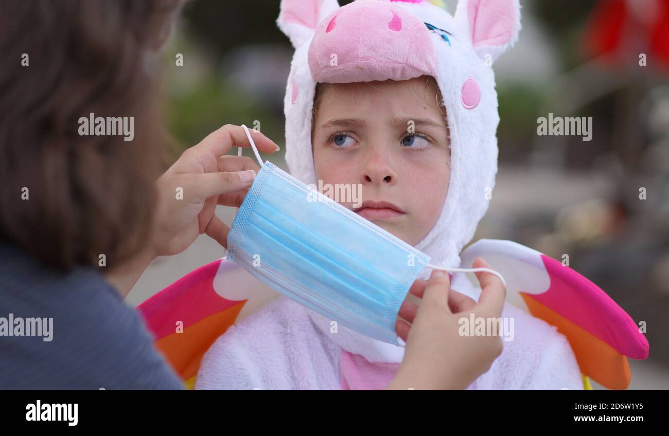 A young girl in a Halloween costume looks very unhappy as her mother puts a face mask on her for her safety and protection in times of Covid-19. Stock Photo