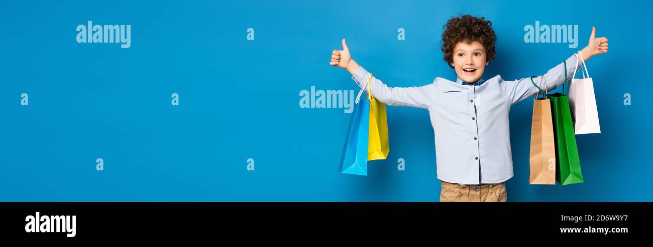 panoramic crop of joyful boy holding shopping bags and showing thumbs up on blue Stock Photo