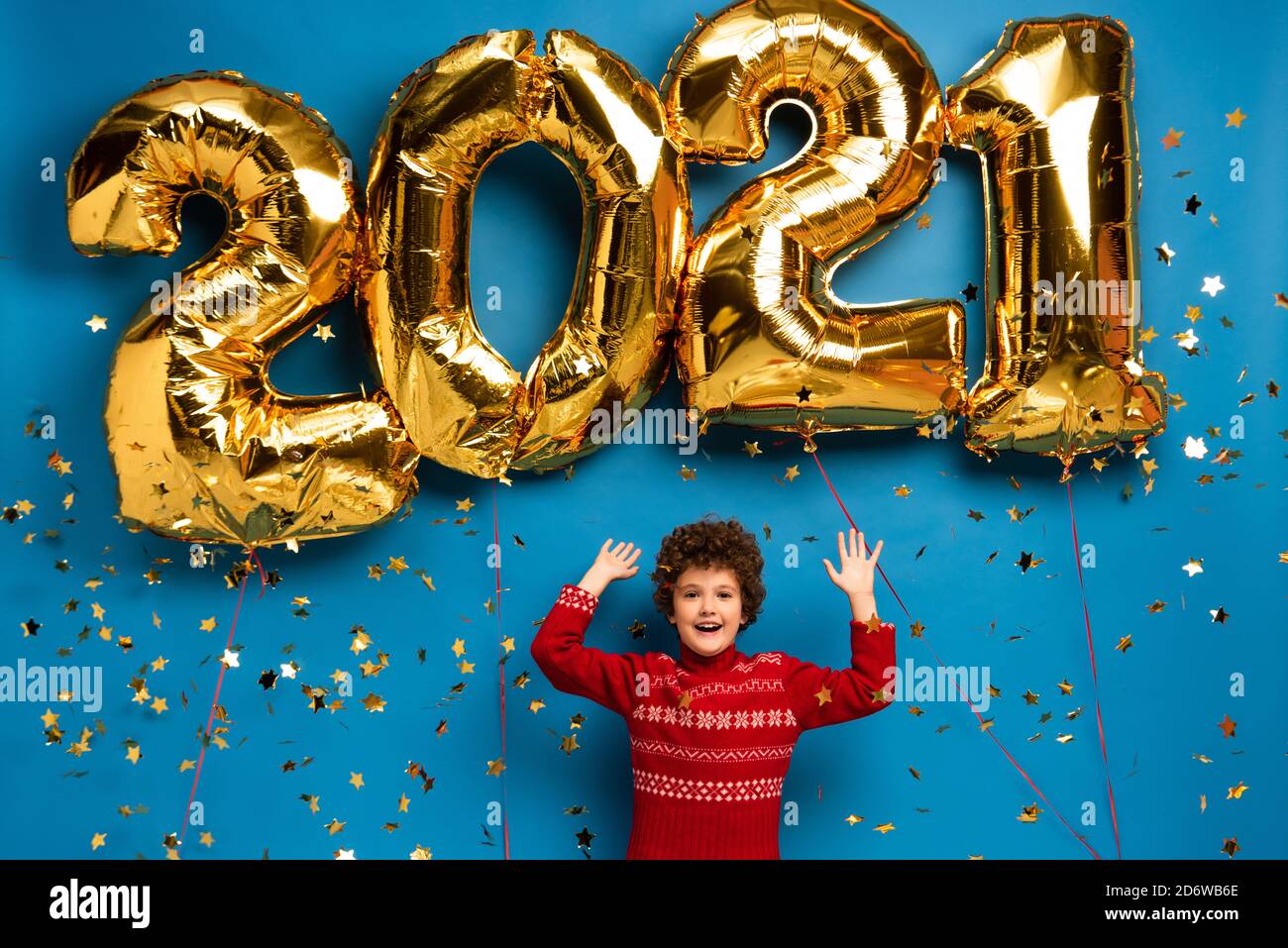 excited boy in red sweater gesturing near golden balloons with 2021 numbers and confetti on blue Stock Photo