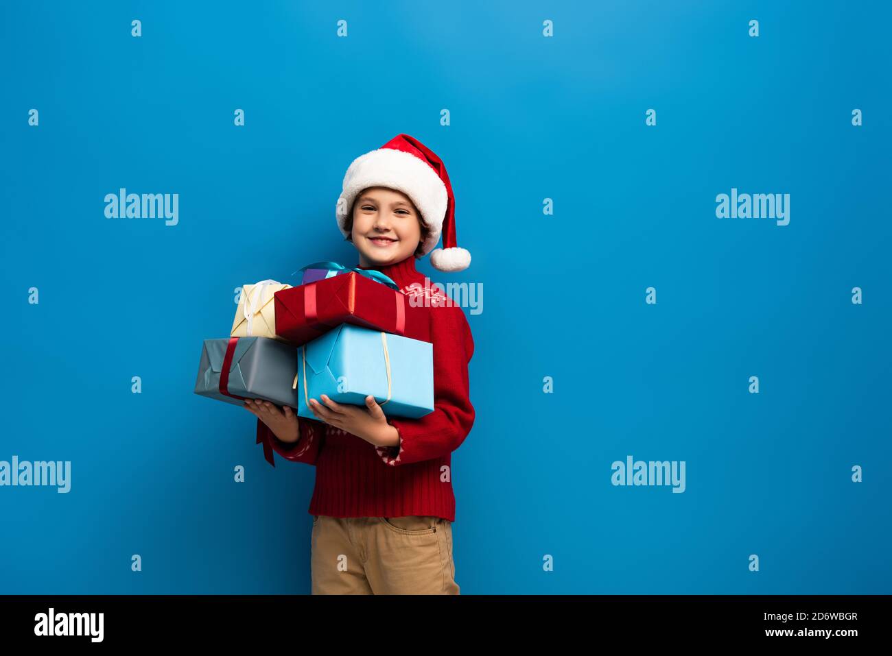 happy boy in santa hat and sweater holding presents on blue Stock Photo