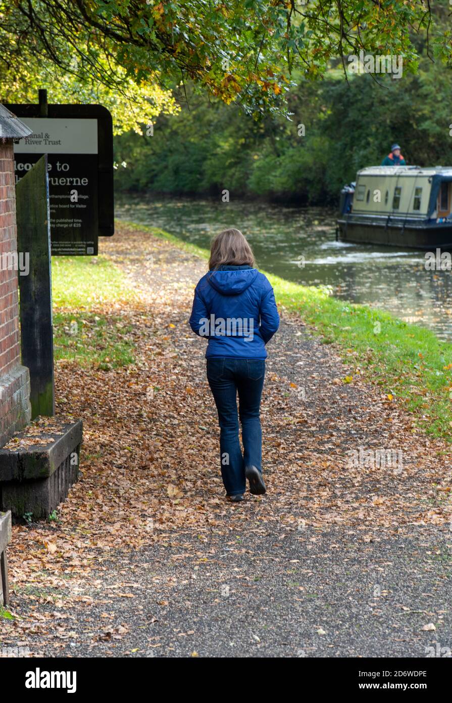 young woman walking along a riverside on a canal towpath with a narrow boat in the background. Stock Photo
