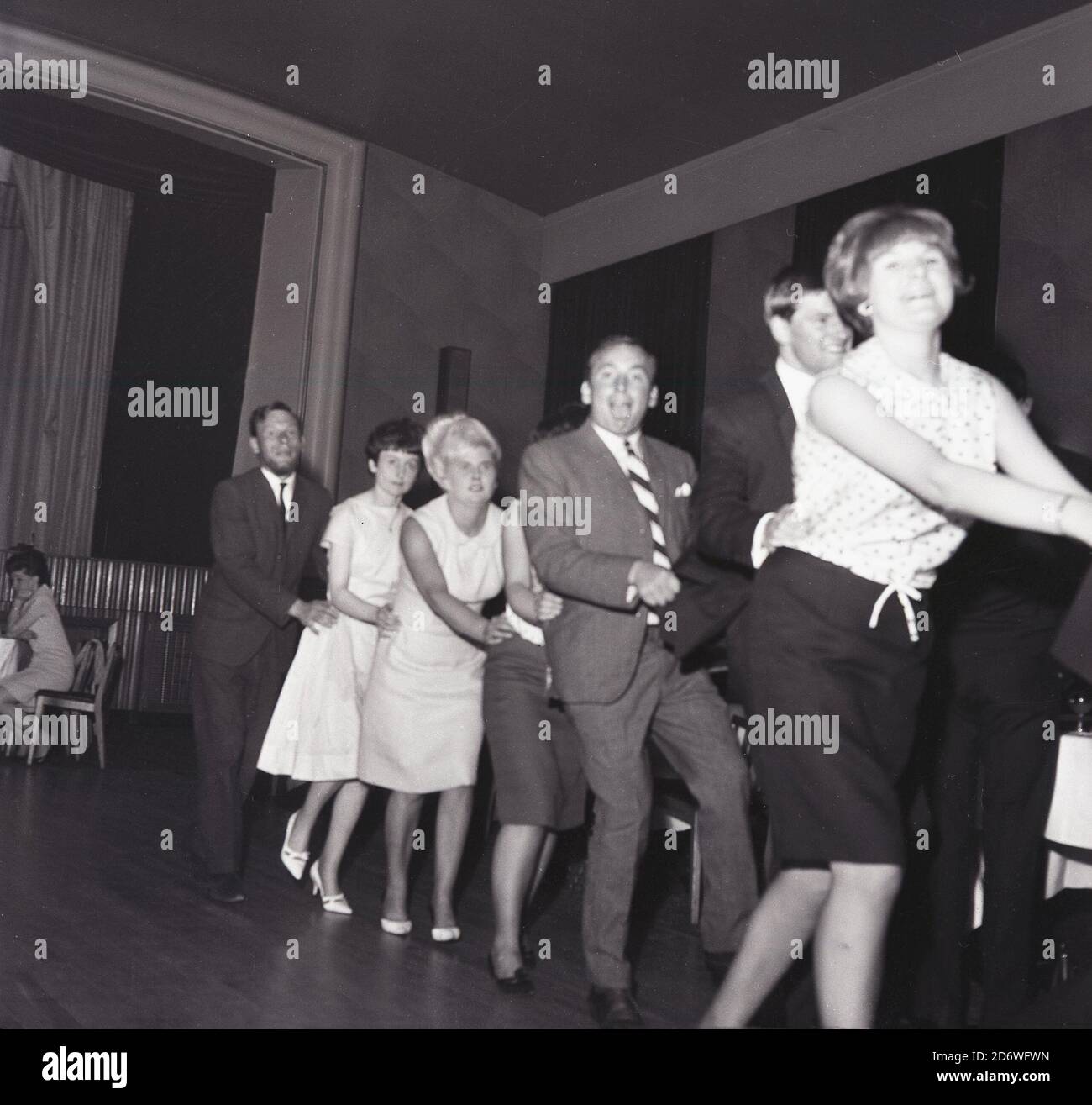 1969, historical, a night  out....evening time and at a party in a large function hall, a number of the guests having fun dancing the conga, a novelty line dance orginating from the Cuban carnival dance and popular in the 1930a and 1950s. Stock Photo
