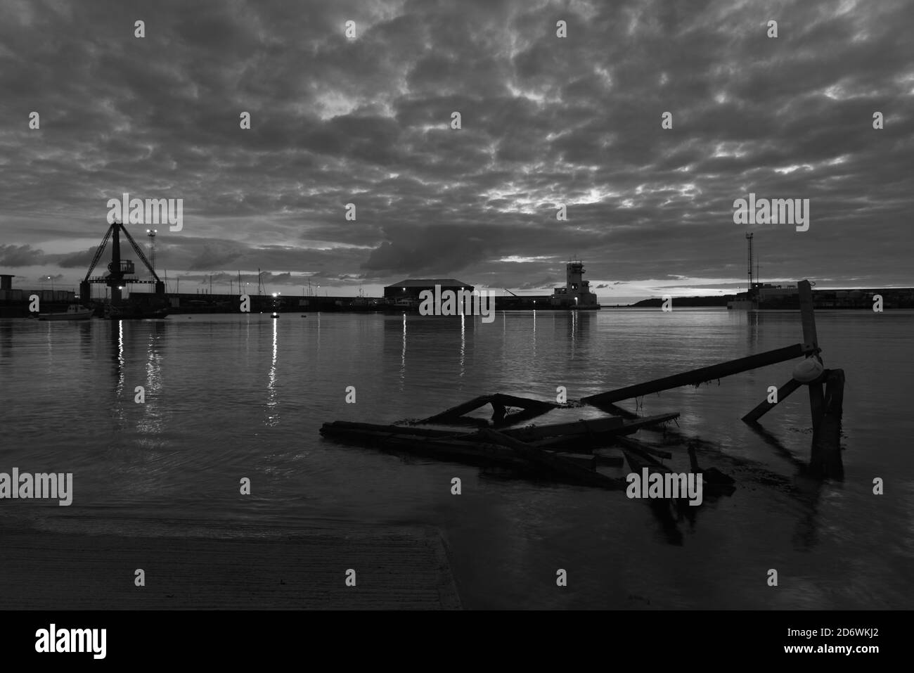 St Helier harbour, Jersey, U.K. B&W image of a calm Autumn high tide, with industrial piers and floating foreground wreckage. Stock Photo