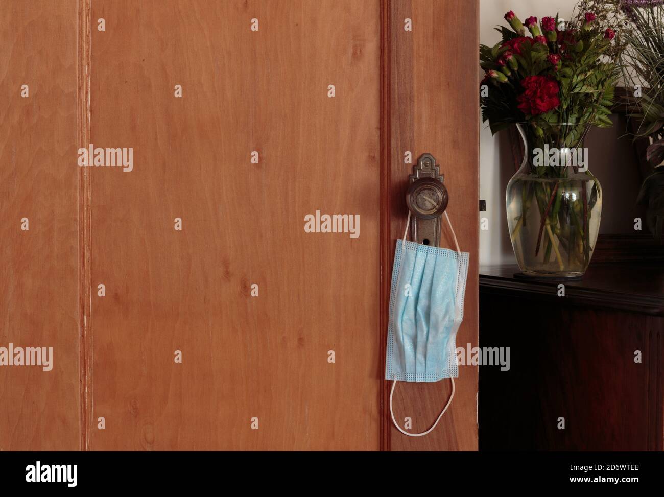 blue face mask hanging from the knob of a wooden door next to a table with red flowers in a vase, theme about face masks becoming a part of daily life Stock Photo