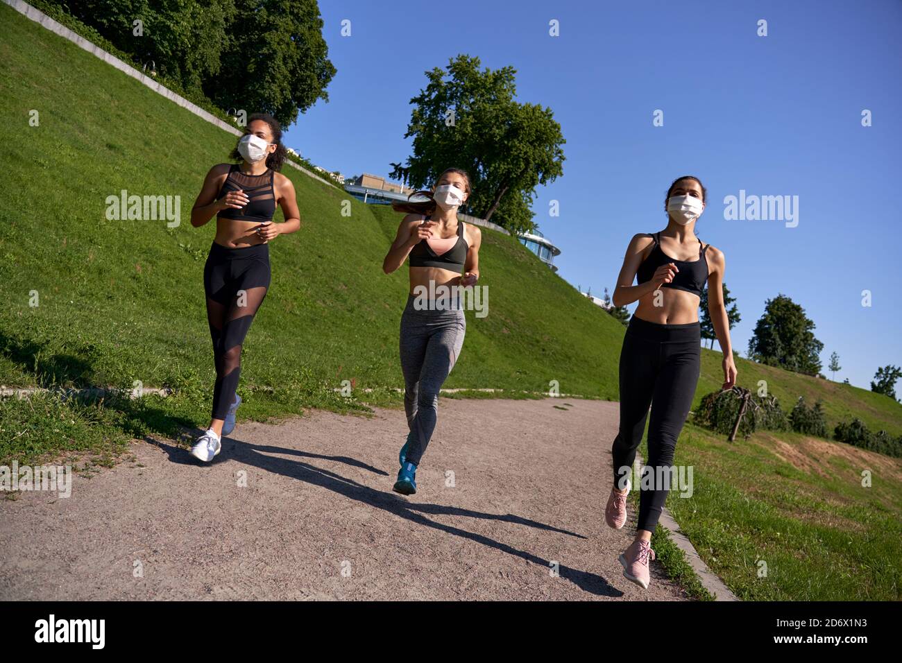 Three multiethnic women group wear face masks run keep social distance outdoor. Stock Photo