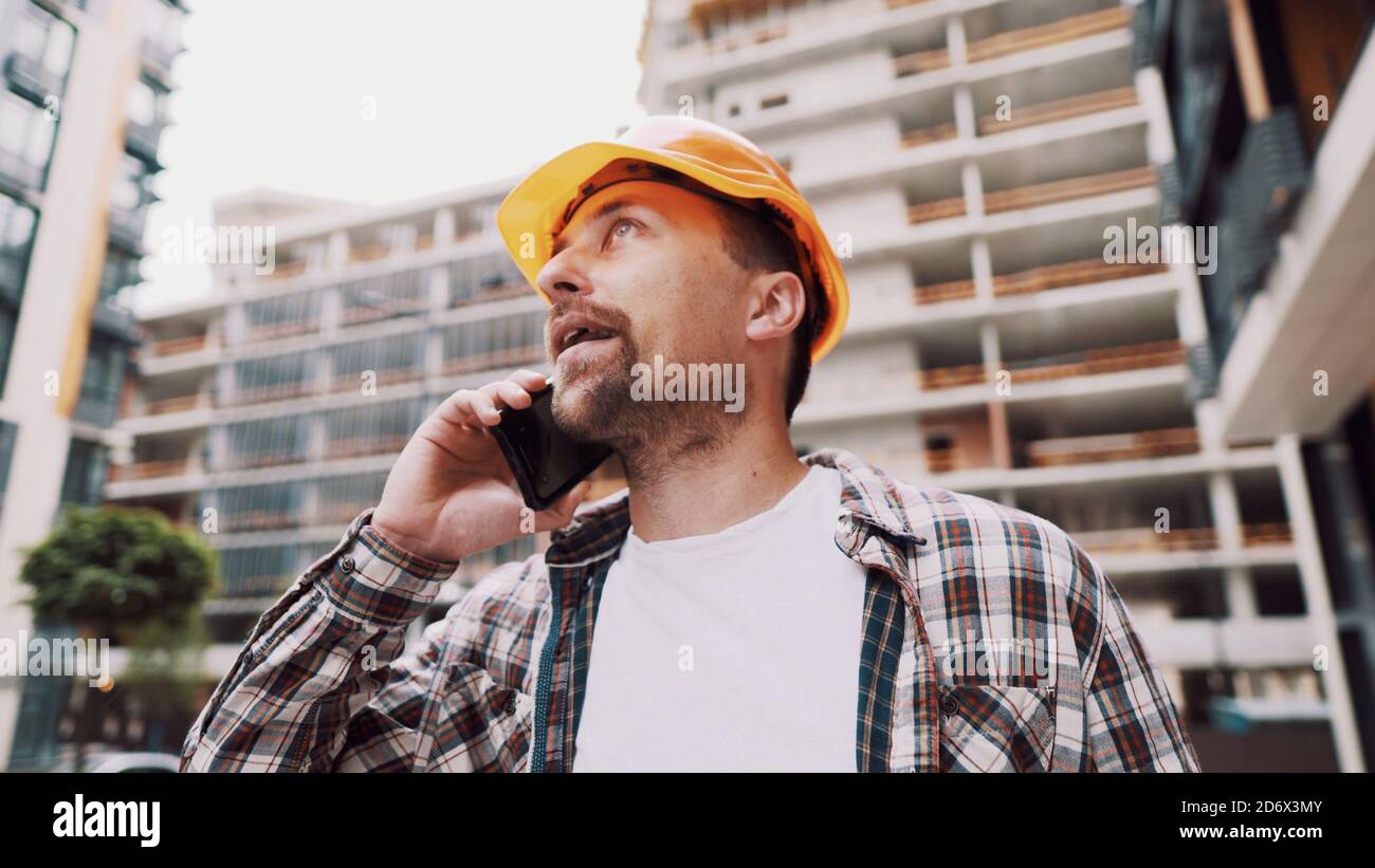 Angry builder, dissatisfied with deadline of work, swears at stress while talking to foreman on phone background of construction site. Wrathful Stock Photo