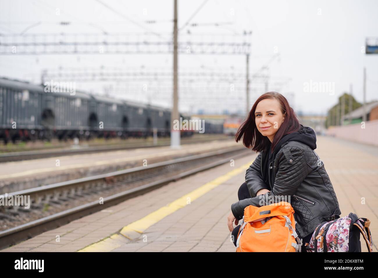 Young woman squats on platform, waiting for train. Female passenger with backpacks sitting on railroad platform in waiting for train ride. Stock Photo