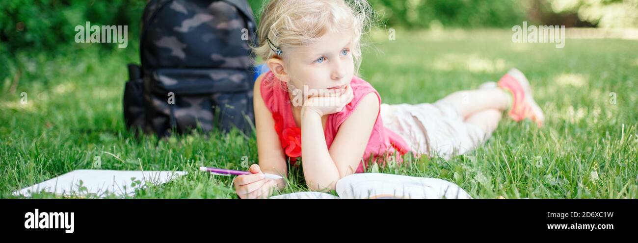 Back to school. Young Caucasian school girl lying in park outdoor doing school homework. Child kid reading book and writing with pencil. Stock Photo