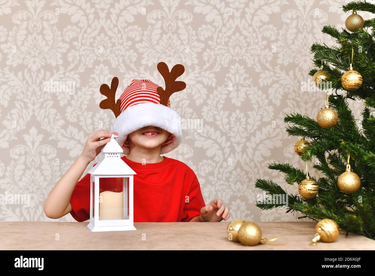 The child sits, near the Christmas tree. Jokingly stretched over her eyes, a cap. Smiles open smile with no one tooth. Stock Photo