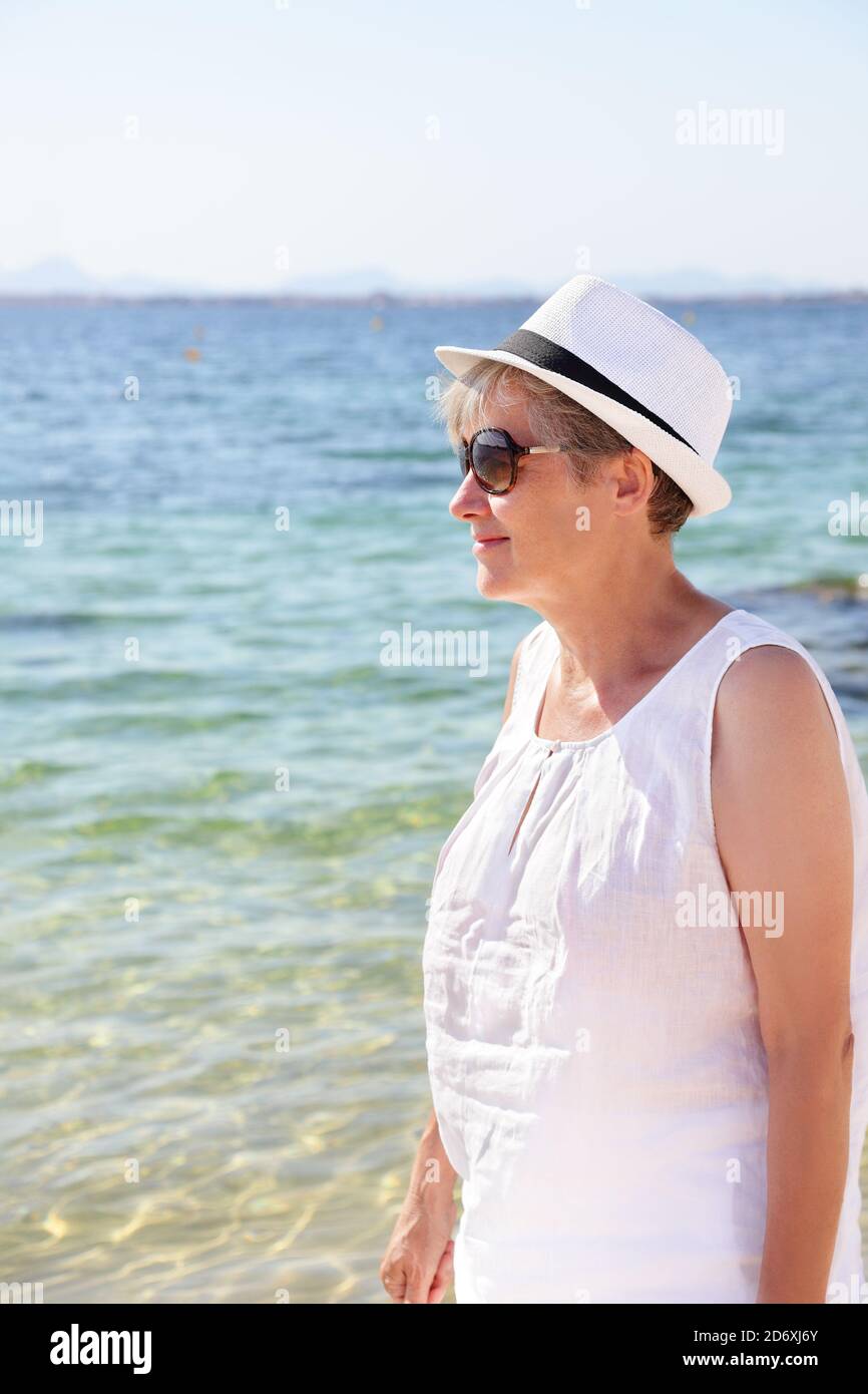 Smiling Senior Woman Walking On Beach By The Sea. Elderly Lady in beachwear wearing white hat and sunglasses. Woman on summer vacation travel holidays Stock Photo