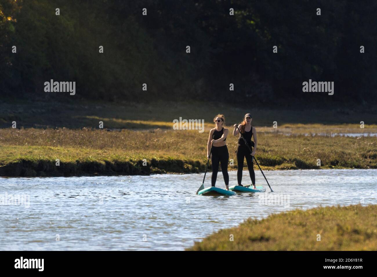 Two female holidaymakers on Stand Up Paddleboards paddling up the Gannel River in early evening light in Newquay in Cornwall. Stock Photo