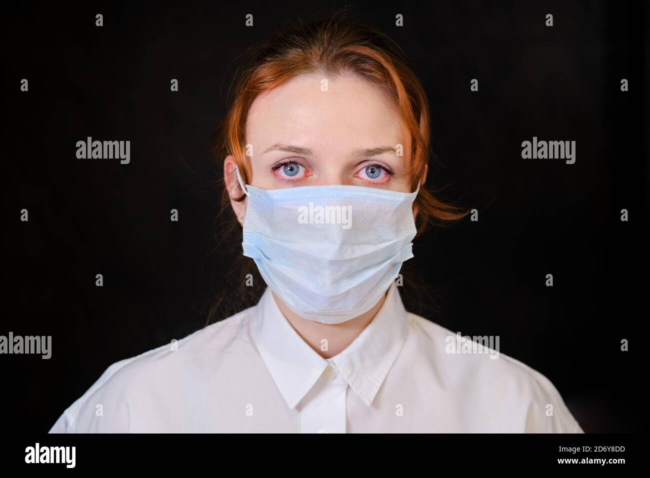 A woman with red eyes in a white shirt and a medical mask on a black background. People caught in isolation due to the flu virus Stock Photo