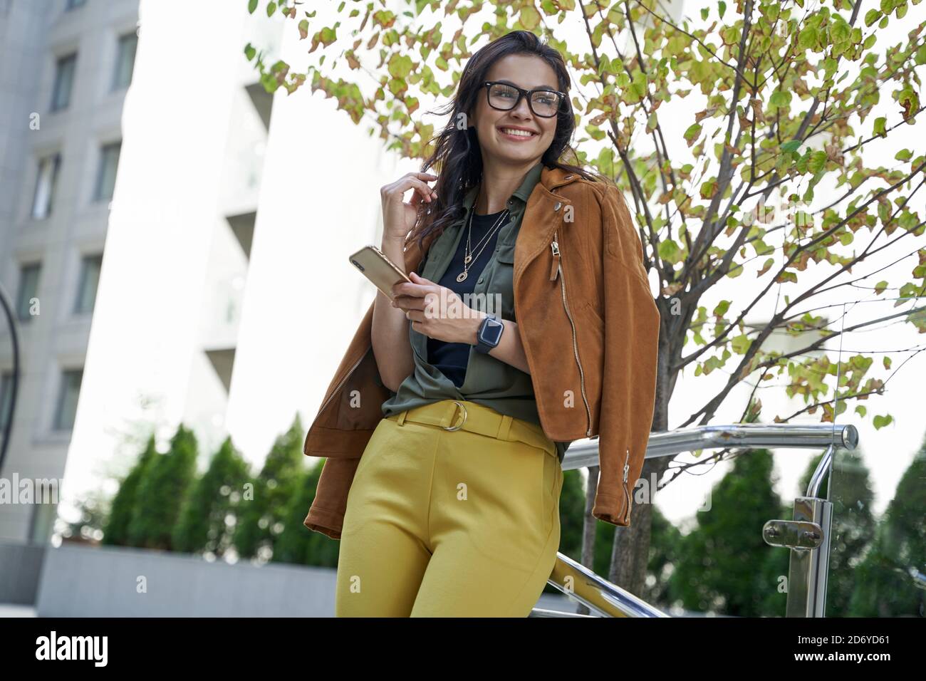 Enjoying autumn day in the city. Young beautiful and stylish woman holding smartphone, looking aside and smiling while walking outdoors. Urban lifestyle concept, people Stock Photo