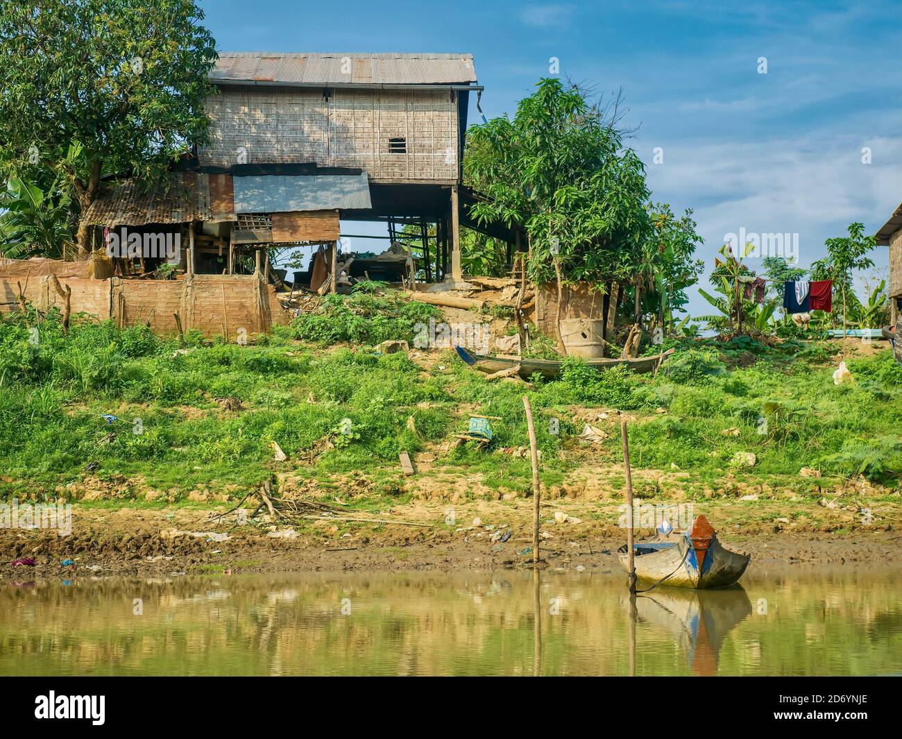 A simple, rural Cambodian house built on wooden stilts, with thatched walls and a metal roof, located beside a river, with a homemade boat in front. Stock Photo